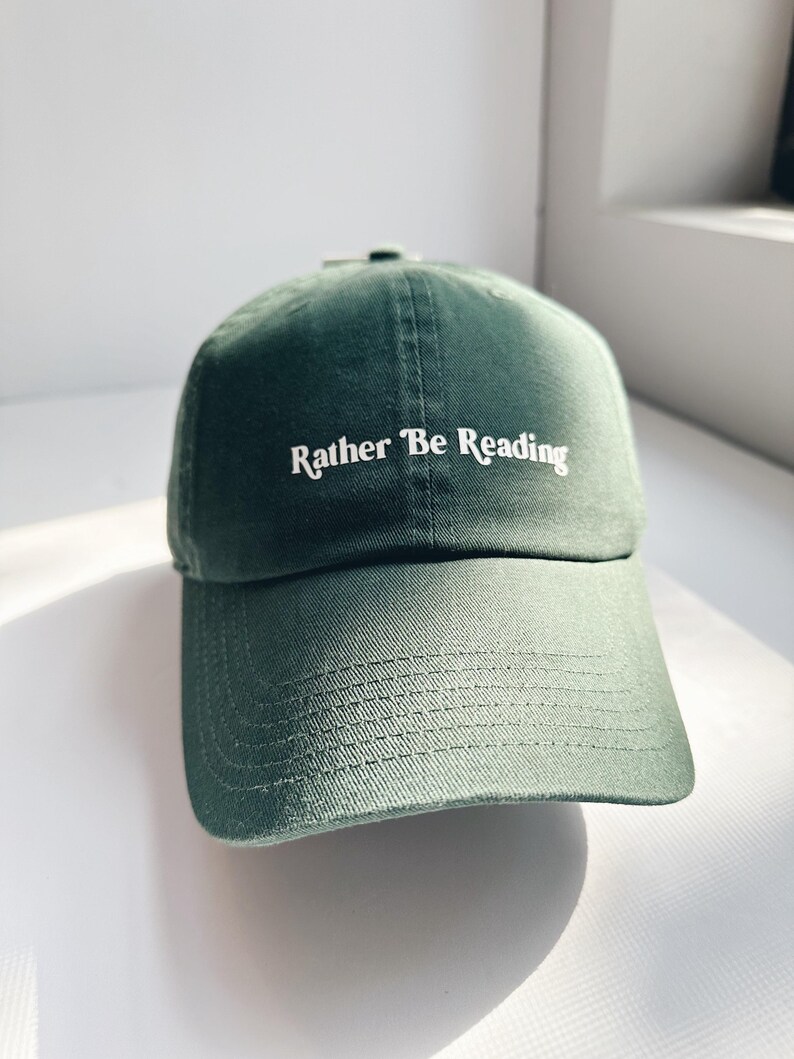 May include: A dark green baseball cap with the text "Rather Be Reading" in white script. The cap is made of a soft fabric and has a curved brim. The hat is a classic style.
