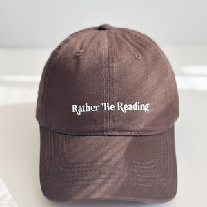 May include: A brown baseball cap with the text "Rather Be Reading" in white. The cap features a curved peak and a rounded crown. The hat is made of a durable fabric, suitable for everyday wear.