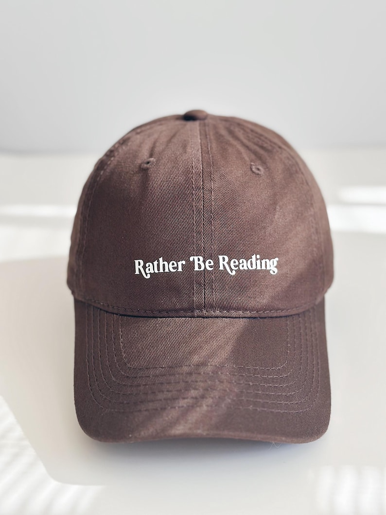 May include: A brown baseball cap with the text "Rather Be Reading" in white. The cap features a curved peak and a rounded crown. The hat is made of a durable fabric, suitable for everyday wear.