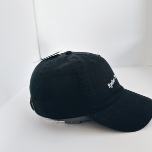 May include: Black baseball cap with the text "Rather Be". The cap has a curved peak and an adjustable strap at the back. The cap is made of a soft, durable fabric.