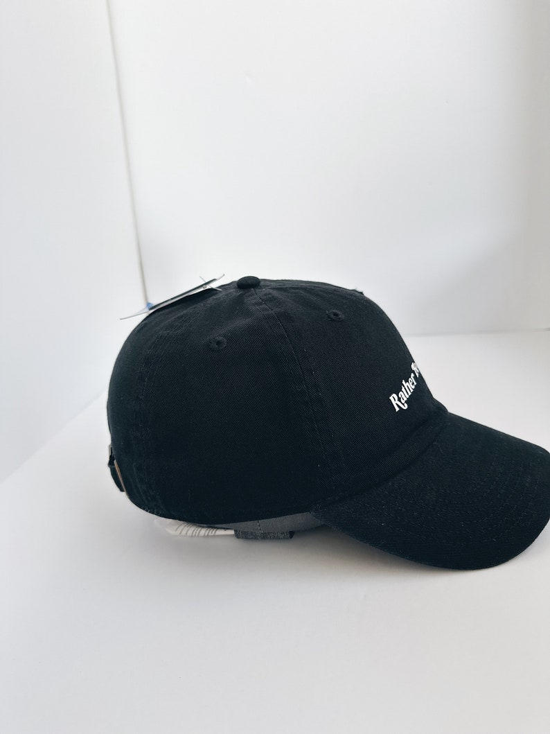 May include: Black baseball cap with the text "Rather Be". The cap has a curved peak and an adjustable strap at the back. The cap is made of a soft, durable fabric.