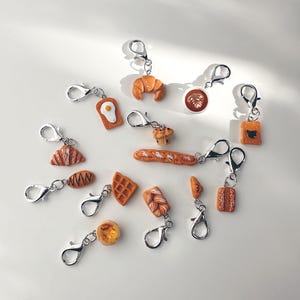 May include: A collection of miniature food-themed keychains. The keychains feature detailed replicas of baked goods like croissants, waffles, and bread, along with a coffee cup and a fried egg. Each keychain has a silver clasp.