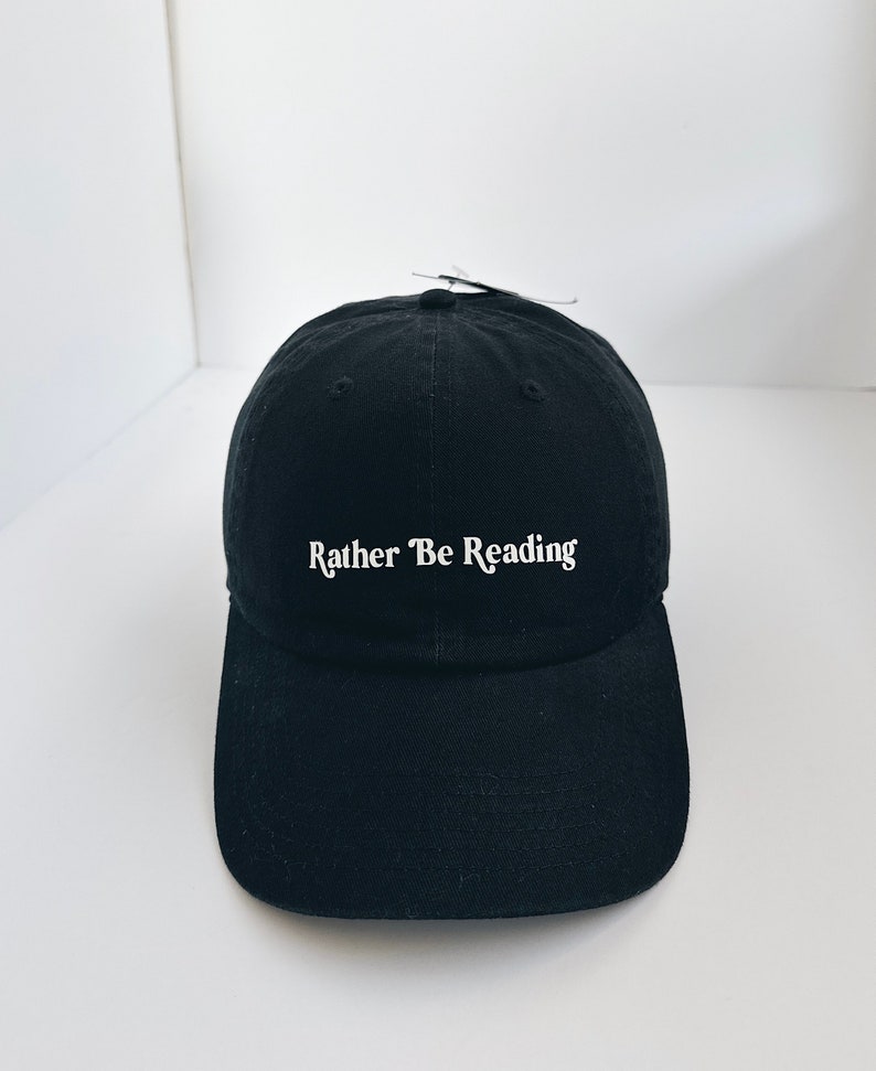 May include: Black baseball cap featuring the white text "Rather Be Reading". The cap has a curved peak and an adjustable fabric strap. A tag is attached to the top of the cap.
