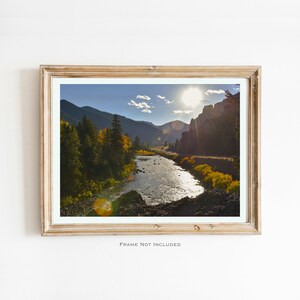 以下が含まれることがあります： 山岳地帯の谷間を流れる川を写した額装された写真。太陽が明るく輝き、水面がキラキラと光っている。川の両岸には緑と黄色の木々が混ざり合って生えている。
