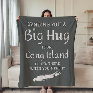 May include: A dark gray blanket with white text that reads "SENDING YOU A Big Hug FROM Long Island SO IT'S THERE WHEN YOU NEED IT." A white outline of Long Island is at the bottom of the blanket. The blanket is held up by a person.