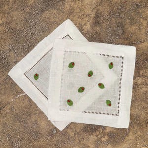 May include: Two white linen cocktail napkins with embroidered green olives and red accents. Each square napkin has a decorative stitched border. The napkins are displayed on a textured, brown surface.
