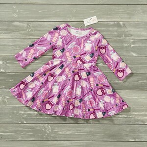 Op de afbeelding: Een roze en witte jurk met lange mouwen met een print van make-up en parfumflessen. De jurk heeft een geruite rok en is perfect voor een klein meisje.