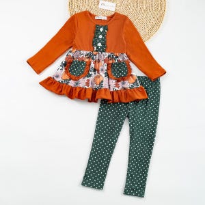 May include: A children's two-piece autumn outfit. The long-sleeved top is burnt orange with a green and white ruffled detail. The skirt has a floral and pumpkin print with green polka dot pockets. The matching leggings are dark green with white polka dots.