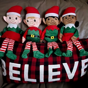 May include: Five plush elf ornaments with red and green hats and outfits, sitting on a red and black plaid pillow with the word "BELIEVE" embroidered in white.