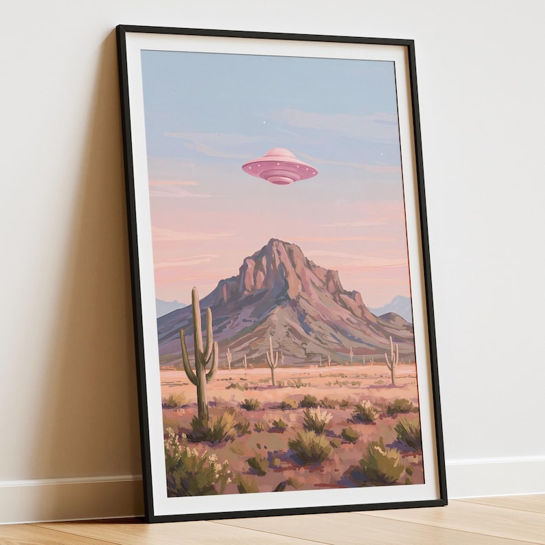 Puede incluir: L&aacute;mina art&iacute;stica enmarcada que representa un paisaje des&eacute;rtico con un OVNI rosa sobre una monta&ntilde;a. La obra de arte muestra cactus, arbustos y un cielo pastel, creando una escena caprichosa y surrealista. La impresi&oacute;n est&aacute; encerrada en un marco negro.