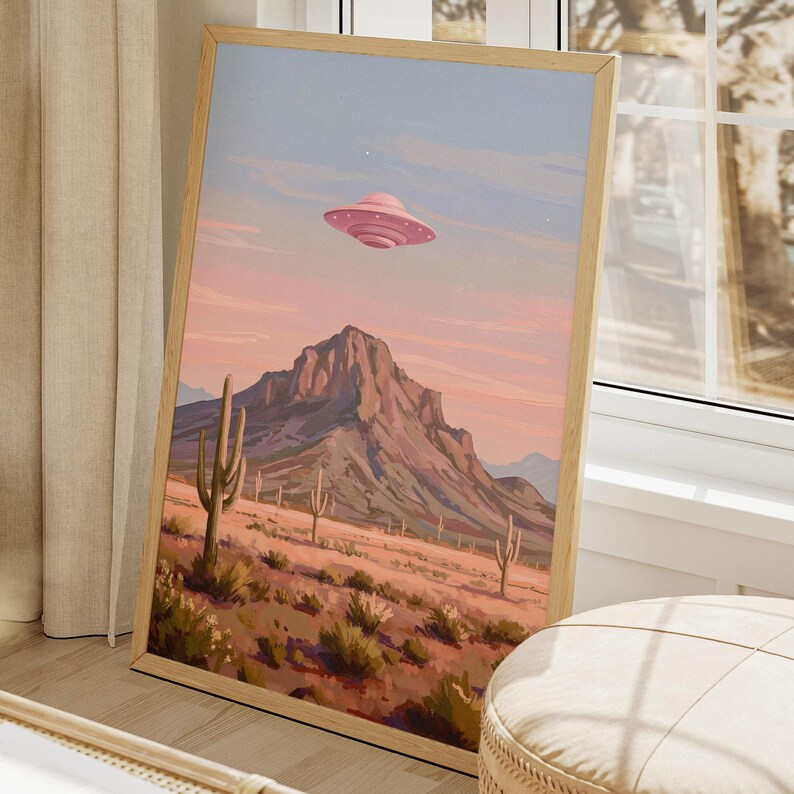 Puede incluir: Una impresi&oacute;n art&iacute;stica enmarcada representa un paisaje des&eacute;rtico bajo un cielo rosa y azul. Un OVNI rosa flota sobre una cordillera y cactus. La obra de arte est&aacute; en un marco de madera, apoyada contra una ventana.