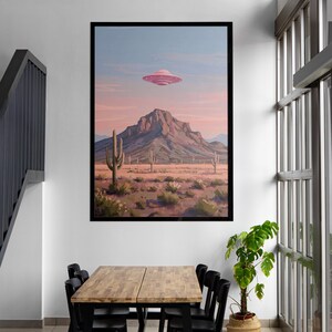 Puede incluir: Una impresi&oacute;n art&iacute;stica enmarcada representa un paisaje des&eacute;rtico con una monta&ntilde;a, cactus y un OVNI rosa en el cielo. La obra de arte es de estilo retro, con una paleta de colores de rosas, naranjas y azules. La impresi&oacute;n se exhibe en una pared blanca sobre una mesa de madera y sillas negras.