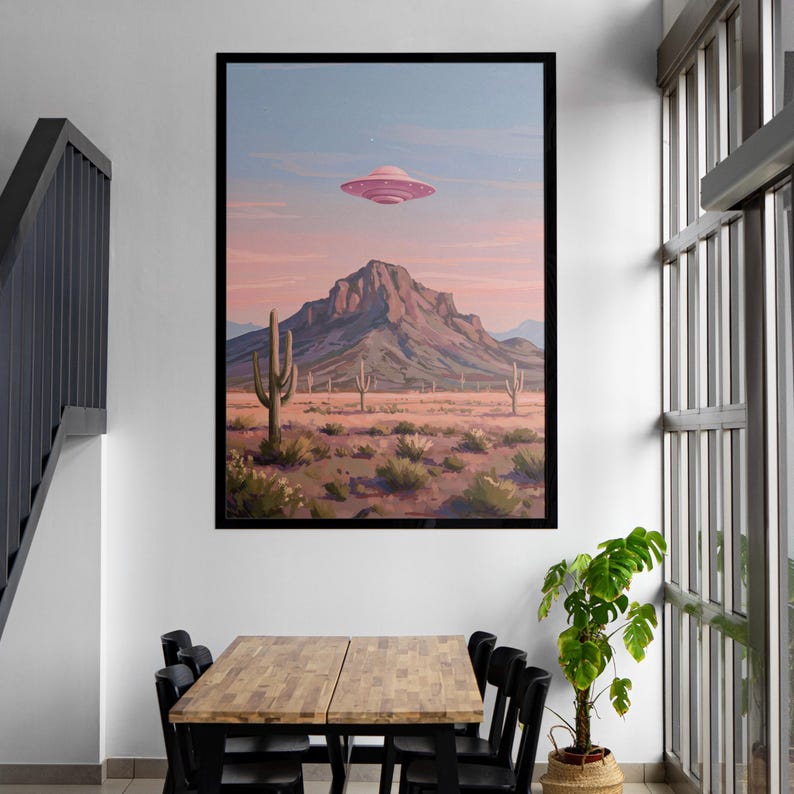 Puede incluir: Una impresi&oacute;n art&iacute;stica enmarcada representa un paisaje des&eacute;rtico con una monta&ntilde;a, cactus y un OVNI rosa en el cielo. La obra de arte es de estilo retro, con una paleta de colores de rosas, naranjas y azules. La impresi&oacute;n se exhibe en una pared blanca sobre una mesa de madera y sillas negras.