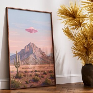 Puede incluir: Una impresi&oacute;n art&iacute;stica enmarcada representa un paisaje des&eacute;rtico con una cordillera, cactus y un OVNI rosa en el cielo. La obra de arte est&aacute; en un marco de madera. La paleta de colores incluye rosa, azul, marr&oacute;n y verde.