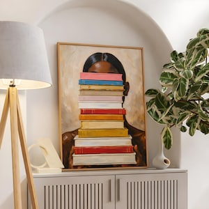 Puede incluir: Una pintura enmarcada representa a una persona con cabello y piel oscuros, con el rostro parcialmente oculto por una pila de libros coloridos. Los libros son en tonos rosa, azul, rojo y amarillo. La obra de arte se exhibe en un mueble blanco.