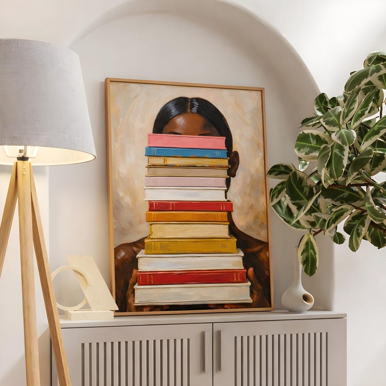 Puede incluir: Una pintura enmarcada representa a una persona con cabello y piel oscuros, con el rostro parcialmente oculto por una pila de libros coloridos. Los libros son en tonos rosa, azul, rojo y amarillo. La obra de arte se exhibe en un mueble blanco.