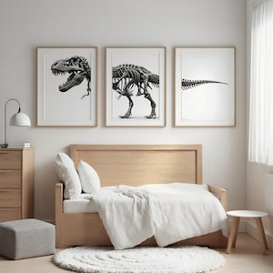May include: Three framed black and white dinosaur skeleton prints. The prints depict a T-Rex skull, a full T-Rex skeleton, and a T-Rex tail. The artwork is displayed above a bed with white bedding.