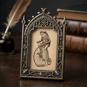 Może przedstawiać: Ozdobna ramka na zdjęcia w kolorze brązu z gotyckim łukiem. Zawiera sepiową ilustrację żaby w garniturze na rowerze. Ozdobne detale obejmują wzory kwiatowe i geometryczne.