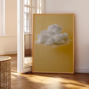 May include: A framed art print featuring a fluffy white cloud against a warm yellow background. The artwork is displayed in a room with natural light and a wooden floor. The frame is a light brown color.
