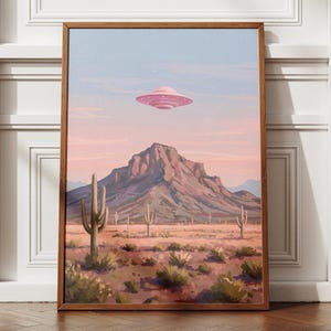 Puede incluir: Una impresi&oacute;n art&iacute;stica enmarcada representa un paisaje des&eacute;rtico con un OVNI rosa flotando sobre una monta&ntilde;a. La obra de arte presenta cactus, arbustos y una paleta de colores pastel rosa, azul y marr&oacute;n. La impresi&oacute;n est&aacute; en un marco de madera.