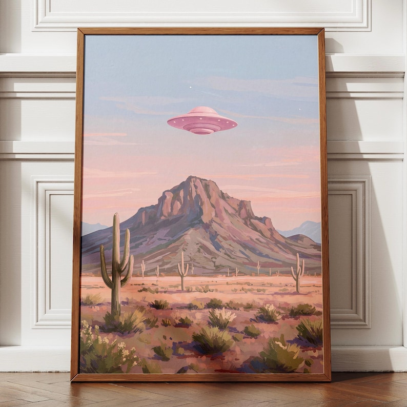 Puede incluir: Una impresi&oacute;n art&iacute;stica enmarcada representa un paisaje des&eacute;rtico con un OVNI rosa flotando sobre una monta&ntilde;a. La obra de arte presenta cactus, arbustos y una paleta de colores pastel rosa, azul y marr&oacute;n. La impresi&oacute;n est&aacute; en un marco de madera.
