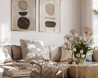 May include: Two framed abstract art prints with beige and brown organic shapes. The artwork hangs above a beige sectional sofa with pillows and a fringed throw. A small gold table holds a vase of white flowers and two brown mugs.