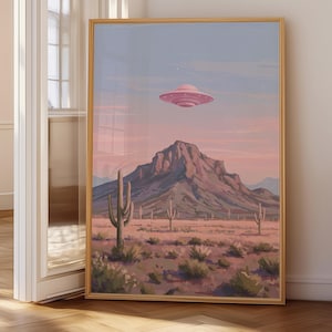 Puede incluir: Una impresi&oacute;n art&iacute;stica enmarcada representa un paisaje des&eacute;rtico bajo un cielo rosa y azul. Un OVNI rosa flota sobre una cordillera, con cactus y plantas del desierto en primer plano. La obra de arte se exhibe en una habitaci&oacute;n.