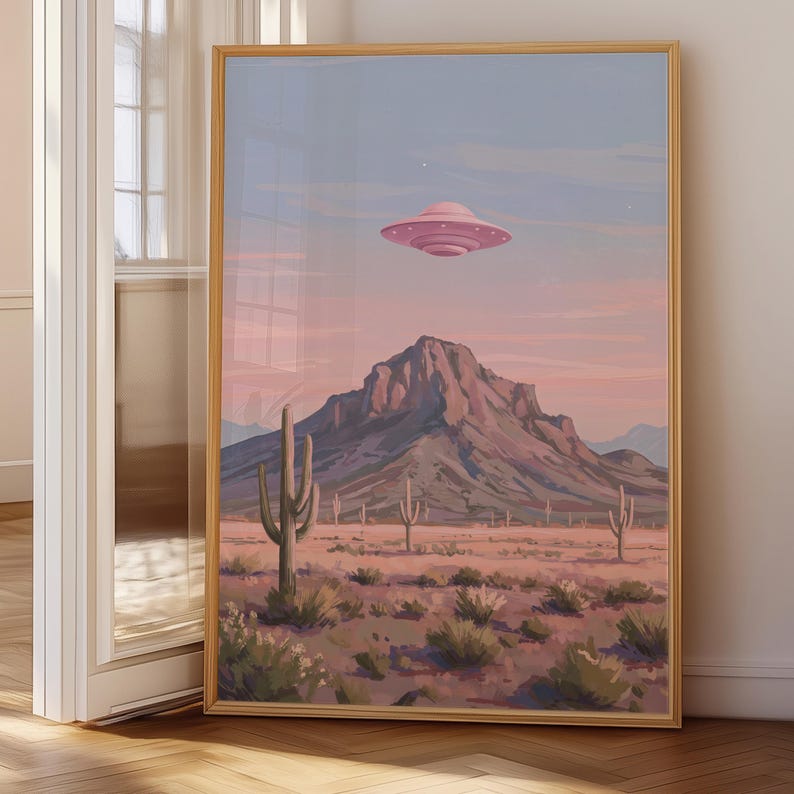 Puede incluir: Una impresi&oacute;n art&iacute;stica enmarcada representa un paisaje des&eacute;rtico bajo un cielo rosa y azul. Un OVNI rosa flota sobre una cordillera, con cactus y plantas del desierto en primer plano. La obra de arte se exhibe en una habitaci&oacute;n.