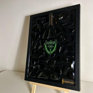 Peut inclure: Une œuvre d'art encadrée représentant une bouteille de champagne. Le cadre noir entoure un fond sombre avec des morceaux de verre noir. La bouteille a une étiquette verte avec du texte et une étoile. L'œuvre est exposée sur un petit chevalet en bois.