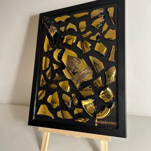 Peut inclure: Une œuvre d'art encadrée représentant une bouteille de champagne noire et des éclats dorés sur fond noir. La bouteille a une étiquette dorée avec un symbole de pique. L'œuvre est présentée sur un petit chevalet en bois.