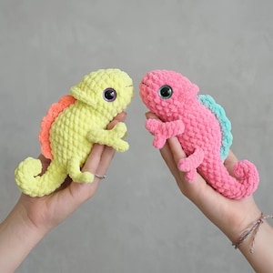 May include: Two handmade crochet chameleons. One is yellow with an orange spine, the other is pink with a light blue spine. Both have large, round, black and purple eyes and are held in hands.