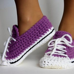Könnte beinhalten: Ein Paar handgefertigte Häkelschuhe im Sneaker-Stil. Die Schuhe sind lila mit weißen Zehen und Sohlen sowie weißen Schnürsenkeln. Die Sohlen sind mit einem schwarzen Streifen umrandet. Die Schuhe sind wie klassische Sneaker gestaltet.