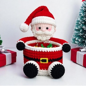 Op de afbeelding: Een gehaakte Santa Claus mand in rood, wit en zwart. De mand heeft de vorm van Santa's lichaam, met een rood pak, zwarte laarzen en een witte baard en muts. De mand is gevuld met kleurrijke snoepjes, perfect voor Kerstmis.