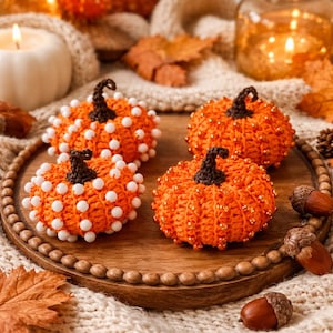 May include: Four handmade orange crochet pumpkins with brown stems and white or gold bead accents sit on a wooden tray. The pumpkins are surrounded by fall-themed decor, including candles and leaves.