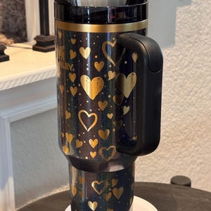 May include: A black and gold insulated travel mug with a handle, decorated with gold hearts on a dark blue surface. The mug has a black lid and a silver straw. The brand name "Stanley" is visible on the side.