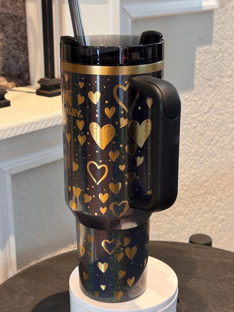 May include: A black and gold insulated travel mug with a handle, decorated with gold hearts on a dark blue surface. The mug has a black lid and a silver straw. The brand name "Stanley" is visible on the side.