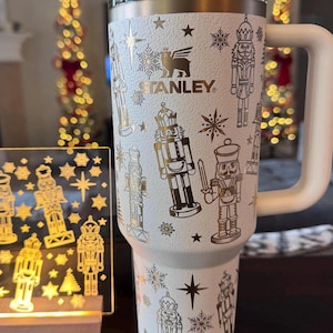 May include: White stainless steel tumbler with a gold lid and handle. The tumbler is decorated with a gold pattern of nutcrackers and snowflakes. The tumbler has the word "STANLEY" printed on it.