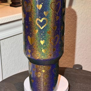 May include: A glittery tumbler with a black lid and gold rim. The tumbler is covered in a pattern of gold hearts on a background of iridescent colours, including blue, green, and purple. The tumbler sits on a white base.