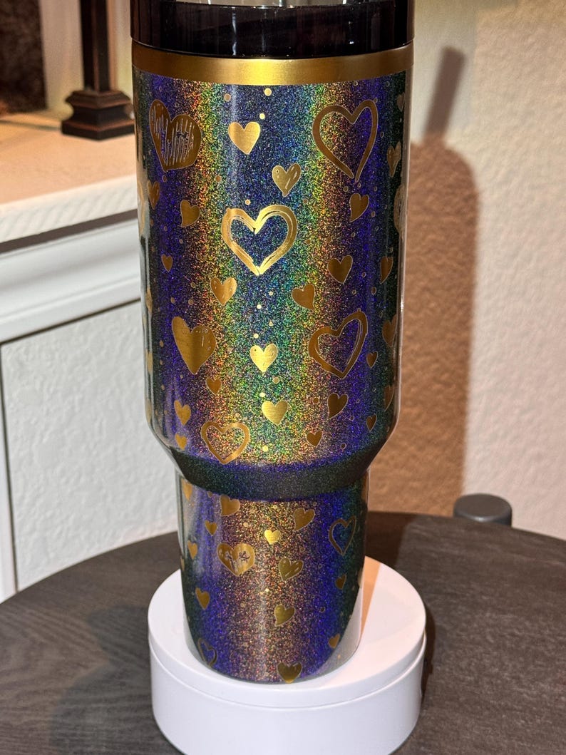 May include: A glittery tumbler with a black lid and gold rim. The tumbler is covered in a pattern of gold hearts on a background of iridescent colours, including blue, green, and purple. The tumbler sits on a white base.