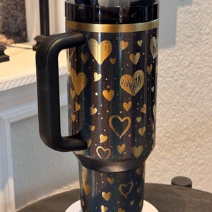 May include: A black travel mug decorated with gold hearts. The mug has a black handle and lid, with a gold band at the top. The design includes gold hearts and dots on a dark background. The mug is suitable for hot or cold drinks.