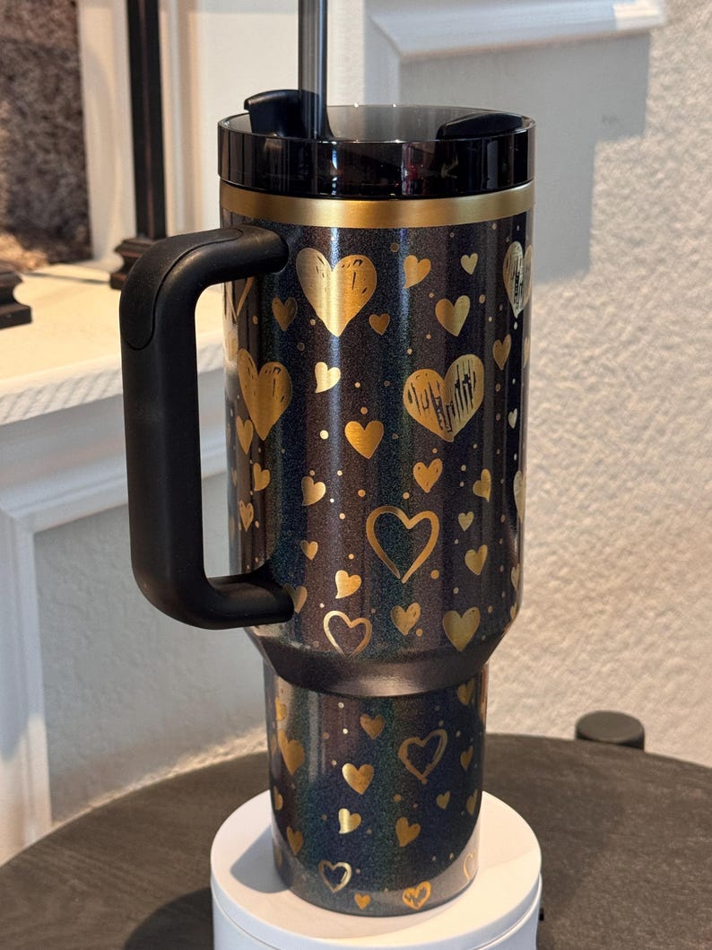 May include: A black travel mug decorated with gold hearts. The mug has a black handle and lid, with a gold band at the top. The design includes gold hearts and dots on a dark background. The mug is suitable for hot or cold drinks.