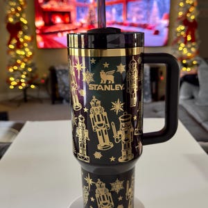 May include: Black and gold insulated tumbler with a handle, featuring a nutcracker and snowflake design. The tumbler has a black lid and a purple straw. The brand name "STANLEY" is visible on the front.