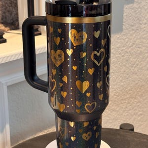 May include: A black and gold insulated travel mug with a handle, decorated with gold hearts and dots. The mug has a black lid and a black straw. The design is glittery and attractive, suitable for hot or cold drinks. The mug is approximately 18 cm tall.