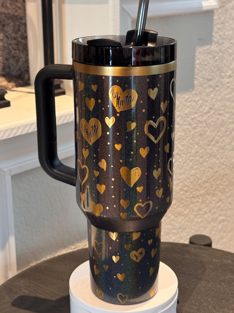 May include: A black and gold insulated travel mug with a handle, decorated with gold hearts and dots. The mug has a black lid and a black straw. The design is glittery and attractive, suitable for hot or cold drinks. The mug is approximately 18 cm tall.