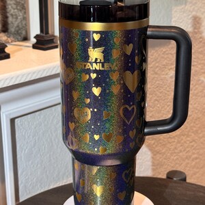 May include: A Stanley travel mug with a black handle and lid. The mug is decorated with gold hearts and the Stanley logo. The background has a glittery, iridescent effect with shades of blue, purple, and gold.