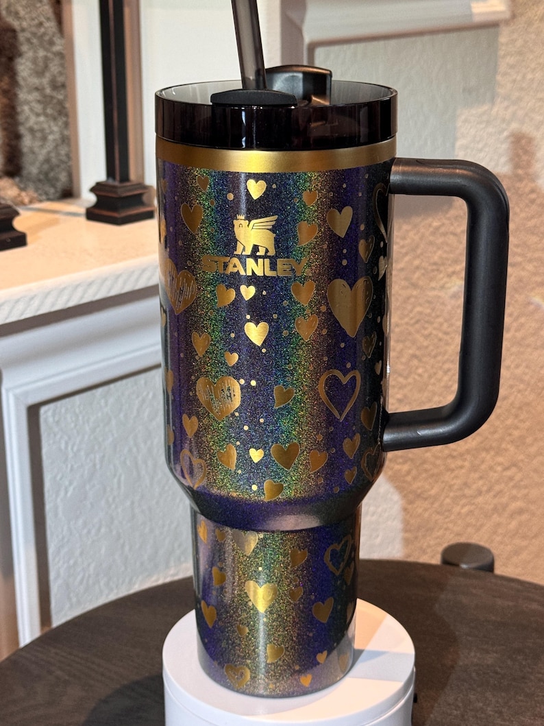 May include: A Stanley travel mug with a black handle and lid. The mug is decorated with gold hearts and the Stanley logo. The background has a glittery, iridescent effect with shades of blue, purple, and gold.