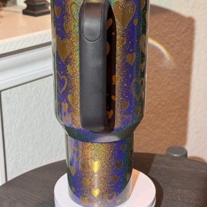May include: A glittery tumbler with a black handle and lid. The tumbler has a blue, green, and gold iridescent background with gold heart shapes. The tumbler is sitting on a white circular base.