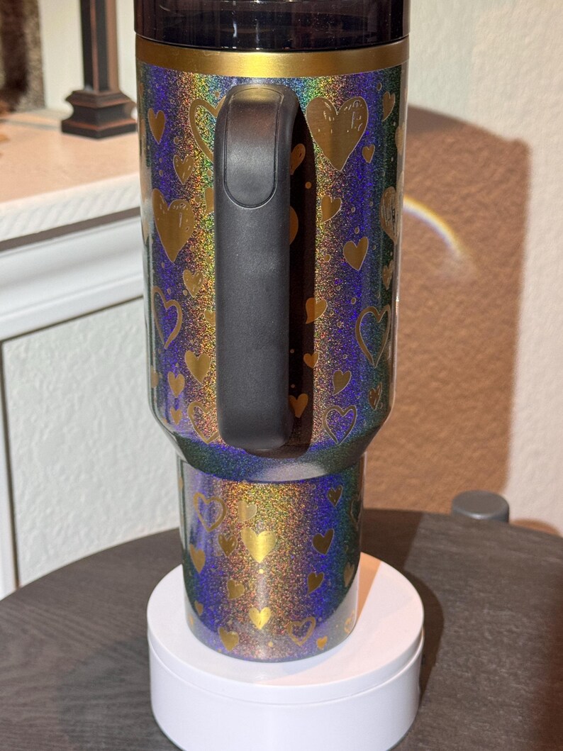 May include: A glittery tumbler with a black handle and lid. The tumbler has a blue, green, and gold iridescent background with gold heart shapes. The tumbler is sitting on a white circular base.