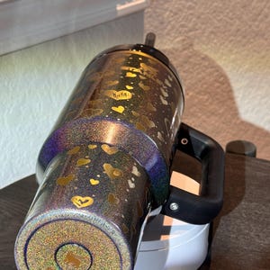 May include: A glittery tumbler with a black handle and a black lid. The tumbler is decorated with gold hearts and a gold logo. The tumbler is approximately 25 cm tall.