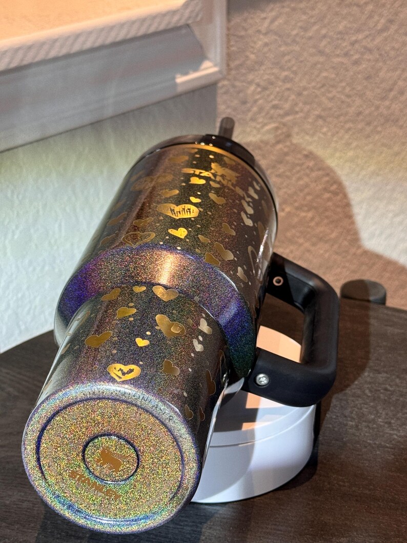 May include: A glittery tumbler with a black handle and a black lid. The tumbler is decorated with gold hearts and a gold logo. The tumbler is approximately 25 cm tall.